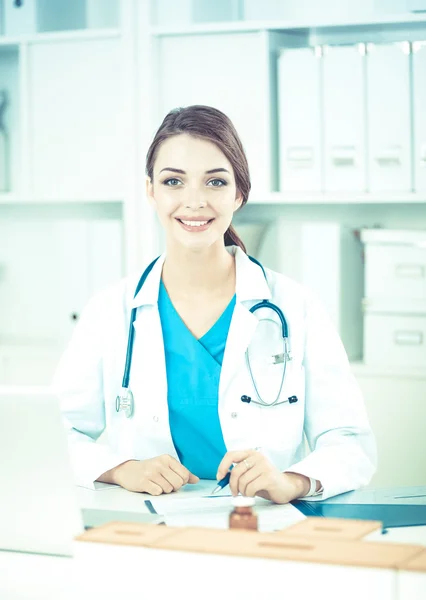 Mooie jonge glimlachende vrouwelijke arts zitten aan het bureau en schrijven. — Stockfoto