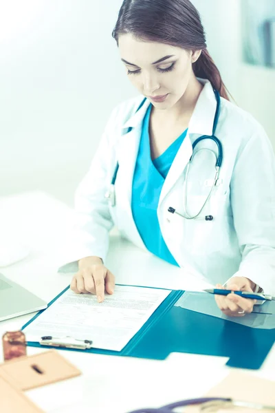 Bonito jovem sorridente médico feminino sentado na mesa e escrevendo. — Fotografia de Stock