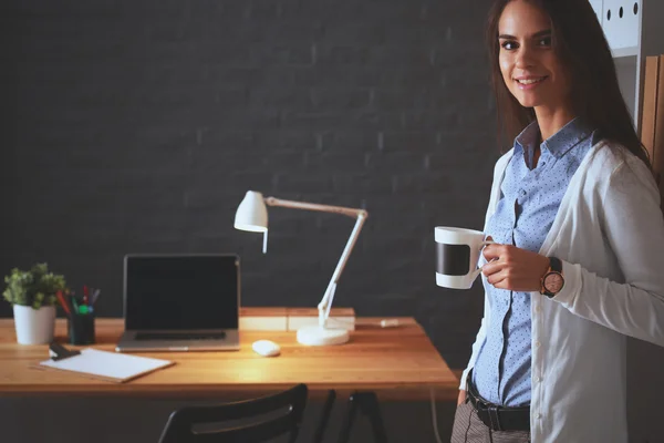 Giovane donna in piedi vicino alla scrivania con computer portatile in possesso di una tazza di caffè — Foto Stock