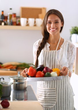 Mutfakta elinde sebzelerle gülümseyen genç bir kadın.