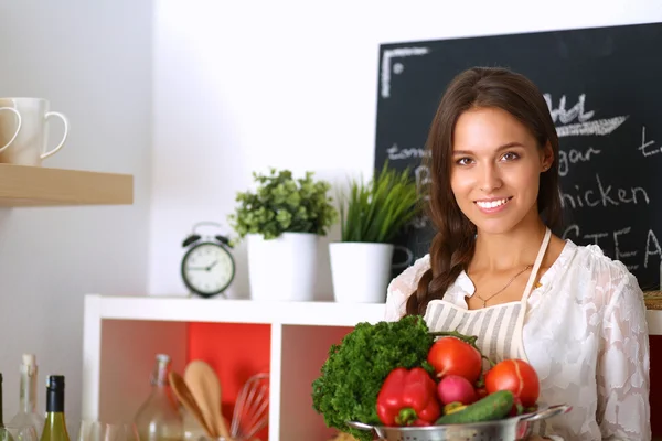 Mujer joven sonriente sosteniendo verduras de pie en la cocina —  Fotos de Stock