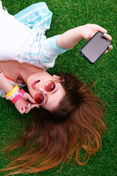 Mooie jonge vrouw selfie maken door haar telefoon liggend in groene gras — Stockfoto