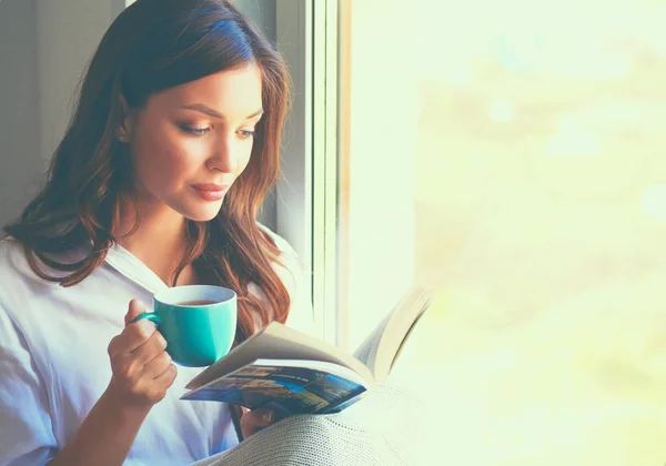Giovane donna a casa seduta vicino alla finestra rilassarsi nel suo soggiorno libro di lettura e bere caffè o tè — Foto Stock