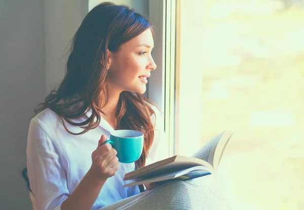 若いです女性で家に座って近く窓リラックスで彼女のリビングルーム読書本と飲料コーヒーや紅茶 — ストック写真