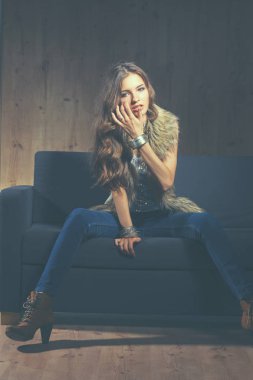 Mavi jeans giydiği siyah koltukta oturan zarif kadın portresi ve kürk yelek