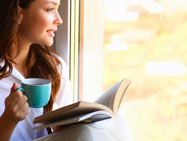 Giovane donna a casa seduta vicino alla finestra rilassarsi nel suo soggiorno libro di lettura e bere caffè o tè — Foto Stock