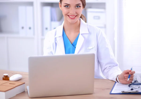 Hermosa joven sonriente doctora sentada en el escritorio y escribiendo —  Fotos de Stock