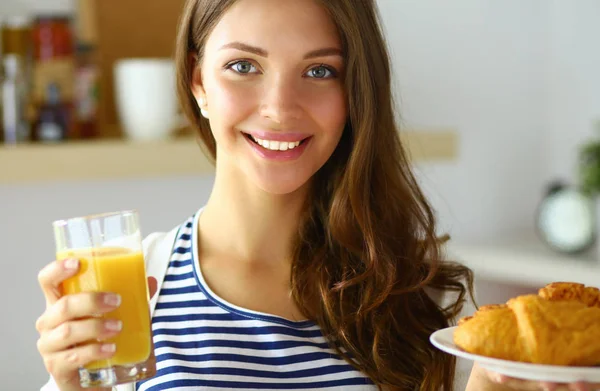 Mujer joven con vaso de jugo y pasteles de pie en la cocina —  Fotos de Stock