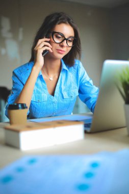 Güzel genç iş kadını ofis masasında oturuyor ve cep telefonuyla konuşuyor.