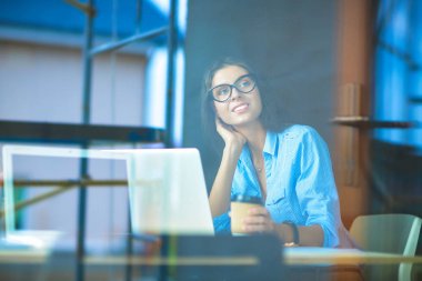 Ofis masasında dizüstü bilgisayarlı genç bir kadın oturuyor, pencereden bakıyor.