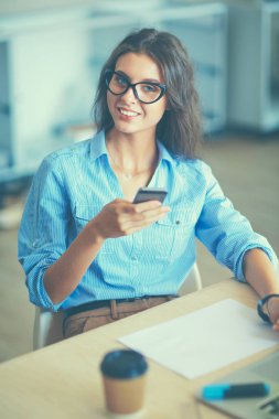 Ofis masasında oturan ve elinde cep telefonu olan genç ve güzel bir iş kadını.