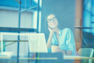 Ofis masasında dizüstü bilgisayarlı genç bir kadın oturuyor, pencereden bakıyor.