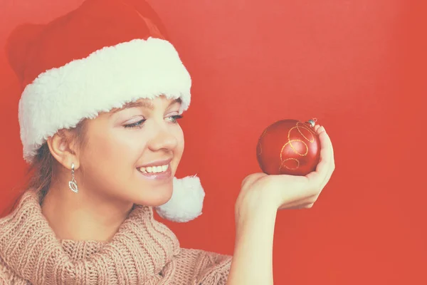 Pretty brunette woman in shirt and santas hat holding red christmas ball — Stock Photo, Image