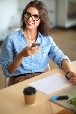 Ofis masasında oturan ve elinde cep telefonu olan genç ve güzel bir iş kadını.