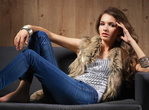 Retrato de una mujer elegante sentada en un sofá negro con pantalones vaqueros azules y chaleco de piel —  Fotos de Stock