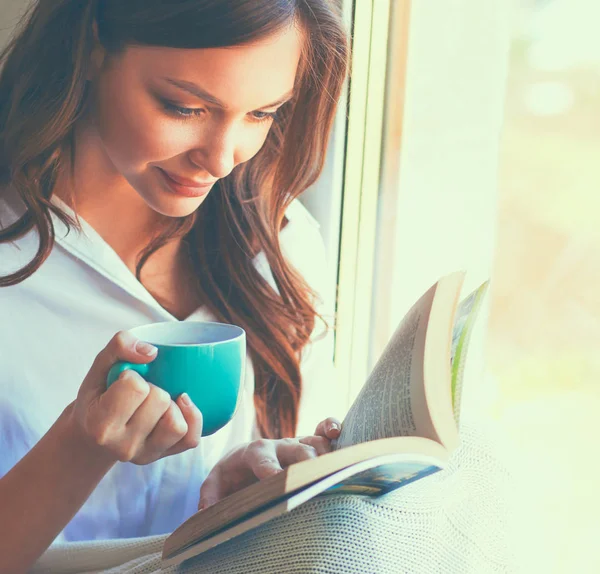 Jovem mulher em casa sentado perto da janela relaxante em sua sala de estar leitura livro e beber café ou chá — Fotografia de Stock