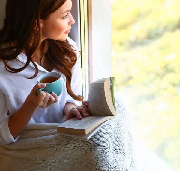 Young woman at home sitting near window relaxing in her living room reading book and drinking coffee or tea — Stock Photo, Image