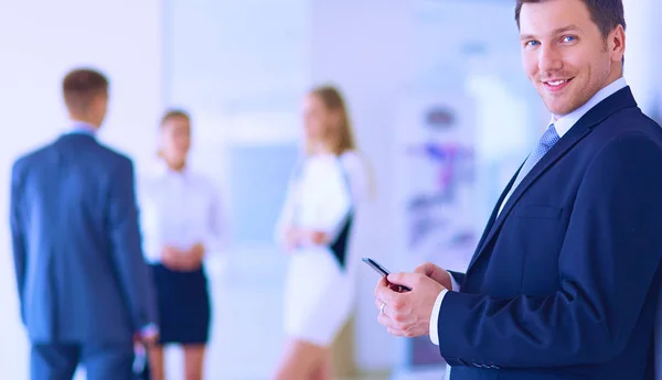 Portrait of young businessman in office with colleagues in the background and using mobile — Stock Photo, Image