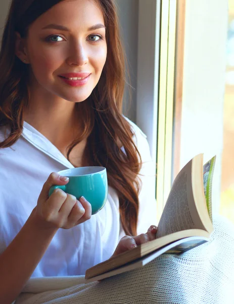 Jovem mulher em casa sentado perto da janela relaxante em sua sala de estar leitura livro e beber café ou chá — Fotografia de Stock