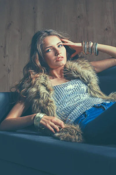 Ritratto di donna elegante seduta sul divano nero con jeans blu e gilet di pelliccia — Foto Stock