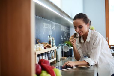 Genç bir kadın mutfakta tablet kullanıyor ve kahve içiyor.