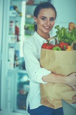 Sebzeli alışveriş poşeti taşıyan genç bir kadın. Mutfakta duruyor.