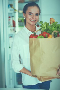 Sebzeli alışveriş poşeti taşıyan genç bir kadın. Mutfakta duruyor.