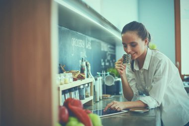 Genç bir kadın mutfakta tablet kullanıyor ve kahve içiyor.