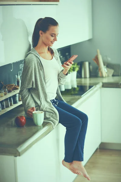 Mujer usando el teléfono móvil sentado en la cocina moderna — Foto de Stock