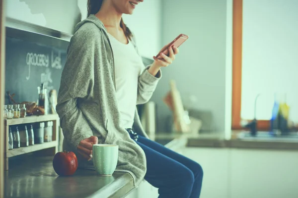 Mujer usando el teléfono móvil sentado en la cocina moderna — Foto de Stock