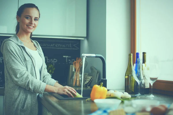 Hermosa joven lavando verduras para ensalada mientras está de pie en la cocina —  Fotos de Stock