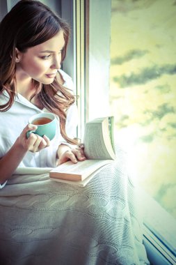 Evde oturan genç bir kadın oturma odasında oturmuş kitap okuyor ve çay ya da kahve içiyor.