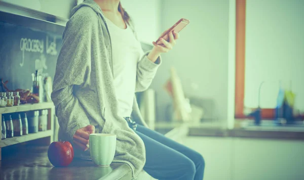 Mujer usando el teléfono móvil sentado en la cocina moderna —  Fotos de Stock