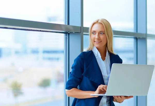 Donna d'affari in piedi contro la finestra dell'ufficio che tiene il computer portatile — Foto Stock