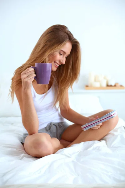 Mooie jonge vrouw lezen van de krant in bed en genieten van een kopje thee — Stockfoto