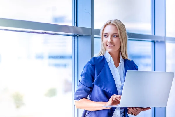 Mujer de negocios de pie contra la ventana de la oficina celebración de ordenador portátil — Foto de Stock