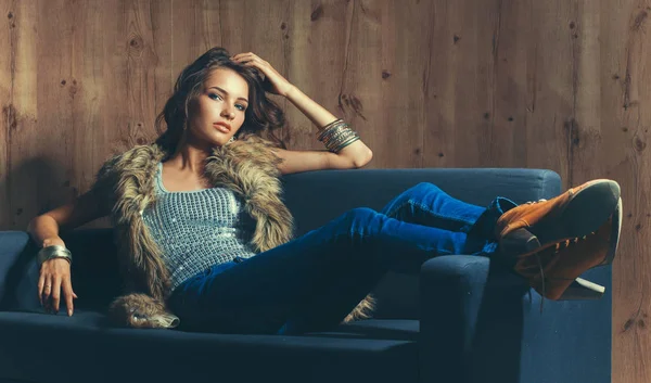 Porträtt av elegant kvinna som sitter på svart soffa klädd i en blå jeans och päls väst — Stockfoto