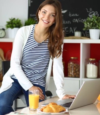 Dizüstü bilgisayar kullanarak ve mutfakta oturmuş çekici genç kadın