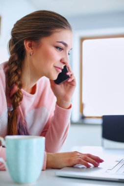 Genç kadın bir dizüstü bilgisayar kullanarak ve cep telefonuyla konuşurken mutfak masasında oturur