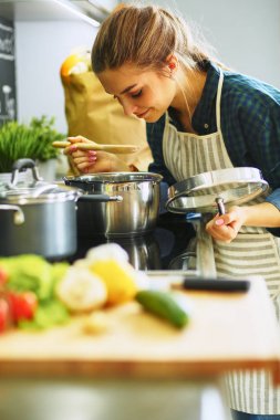 Genç bir kadın mutfağında yemek pişiriyor sobanın yanında duruyor.