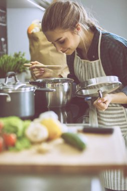 Genç bir kadın mutfağında yemek pişiriyor sobanın yanında duruyor.