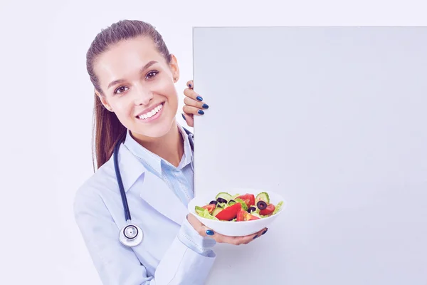 Retrato de uma mulher bonita médico segurando uma placa com legumes frescos em pé perto de branco — Fotografia de Stock