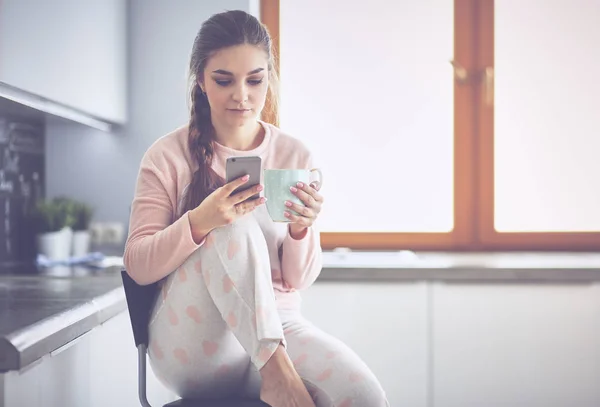 Mujer usando el teléfono móvil sentado en la cocina moderna — Foto de Stock