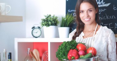 Mutfakta elinde sebzelerle gülümseyen genç bir kadın.