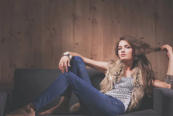 Retrato de una mujer elegante sentada en un sofá negro con pantalones vaqueros azules y chaleco de piel — Foto de Stock