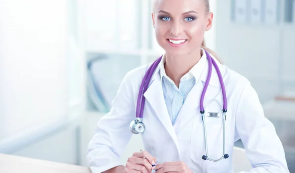 Hermosa joven sonriente doctora sentada en el escritorio y escribiendo. — Foto de Stock