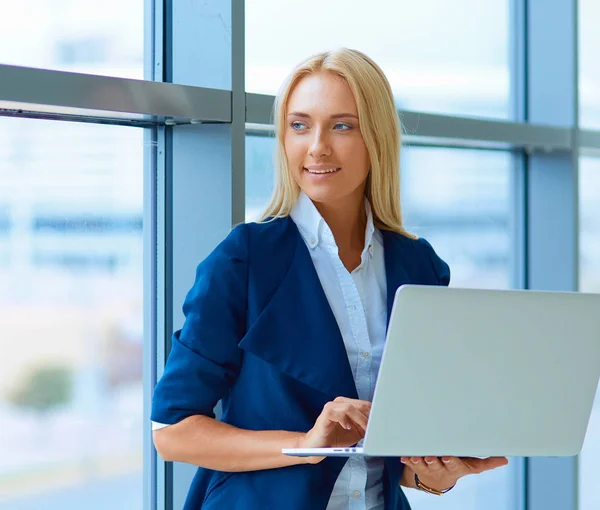 Donna d'affari in piedi contro la finestra dell'ufficio che tiene il computer portatile — Foto Stock