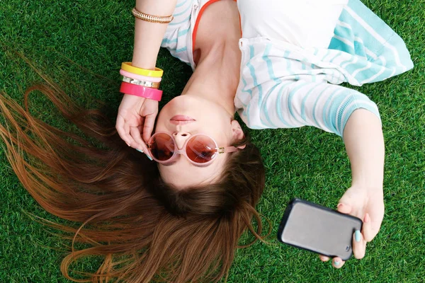 Sorrindo jovem deitada na grama e fazendo uma selfie — Fotografia de Stock