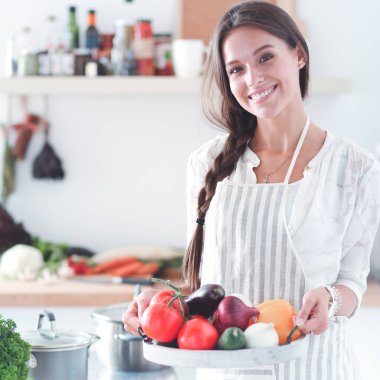 Mutfakta elinde sebzelerle gülümseyen genç bir kadın.