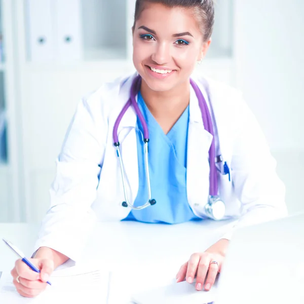 Bonito jovem sorridente médico feminino sentado na mesa e escrevendo. — Fotografia de Stock
